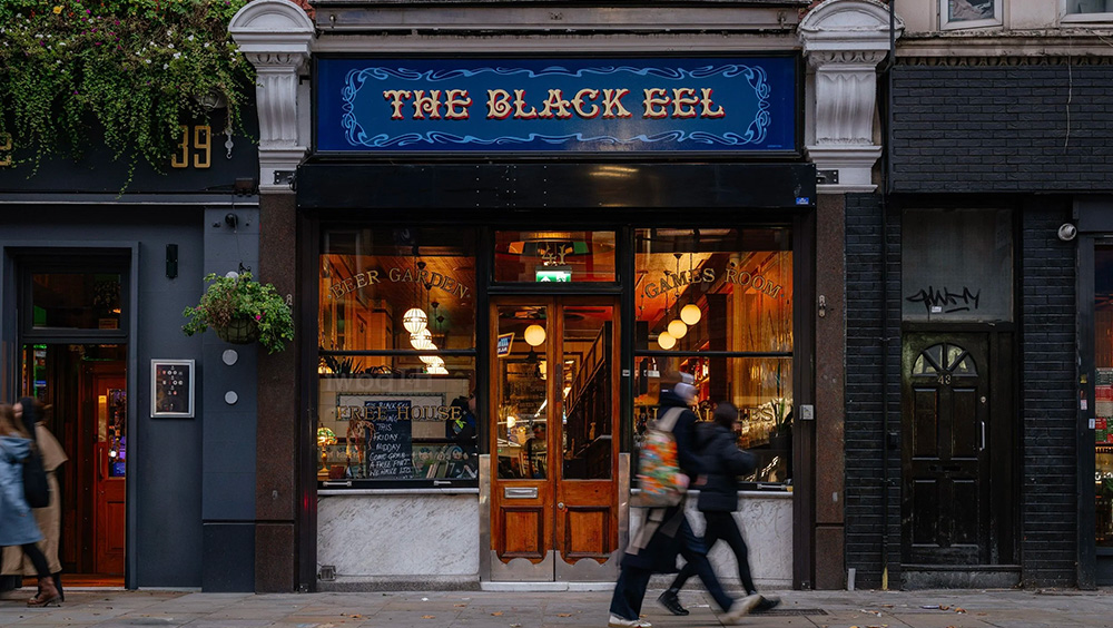 External shot of The Black Eel, with blue signage and gold lettering. Warm inviting amber glow in the windows, with a panelled door