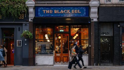 External shot of The Black Eel, with blue signage and gold lettering. Warm inviting amber glow in the windows, with a panelled door